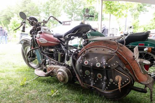 A vintage motorcycle with a rusted body and unique leather saddlebags, parked on green grass under a tent.