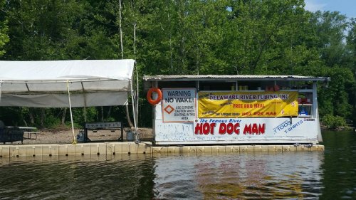 A riverside vendor stand with a "Hot Dog Man" sign, offering tubing services and free BBQ meals, surrounded by trees.