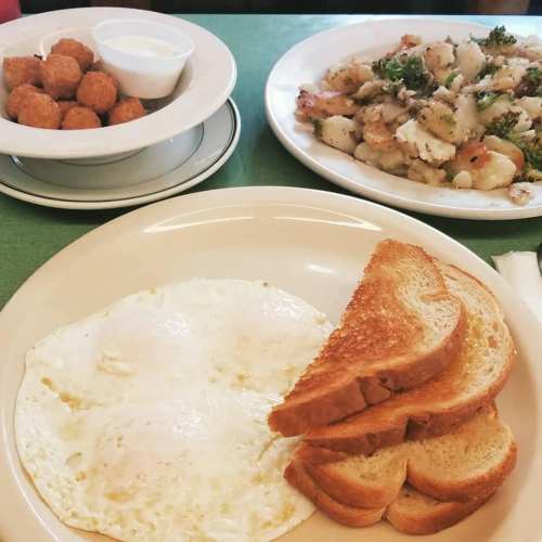 A plate with two fried eggs, toast, and a side of hash browns, alongside a bowl of tater tots and dipping sauce.