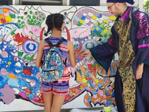 A child in colorful attire paints on a vibrant mural with a man in a robe, both engaged in a creative activity.