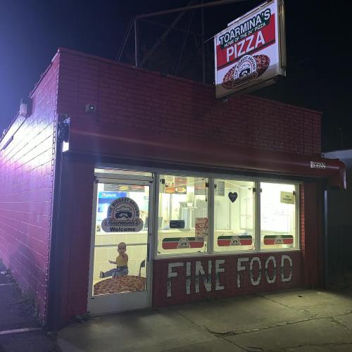 A brightly lit pizza shop at night, with a child sitting inside and a sign reading "Fine Food" on the wall.