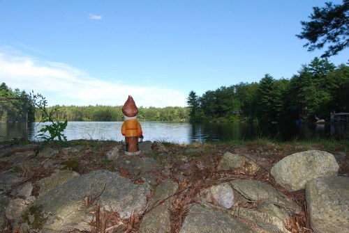 A gnome statue stands on rocks by a serene lake, surrounded by trees under a clear blue sky.