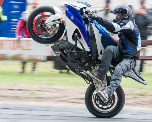 A motorcyclist performs a wheelie on a blue and white bike at a motorsport event, surrounded by spectators.