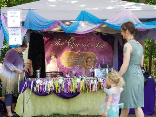 A colorful booth named "The Queen's Ink" with two women and a child, decorated with ribbons and a vibrant backdrop.