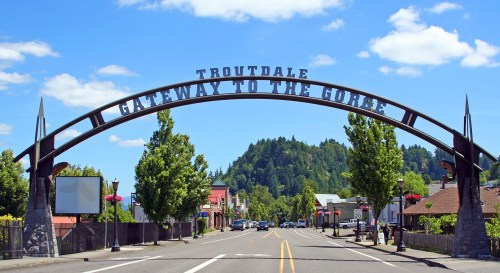 Archway sign reading "Troutdale Gateway to the Gorge" over a tree-lined street with blue skies and mountains in the background.