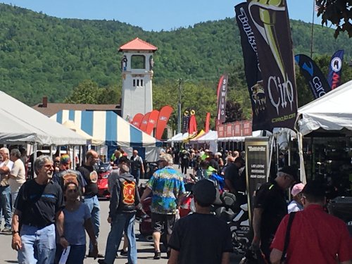 A bustling outdoor event with people browsing vendor tents, surrounded by mountains and colorful flags.
