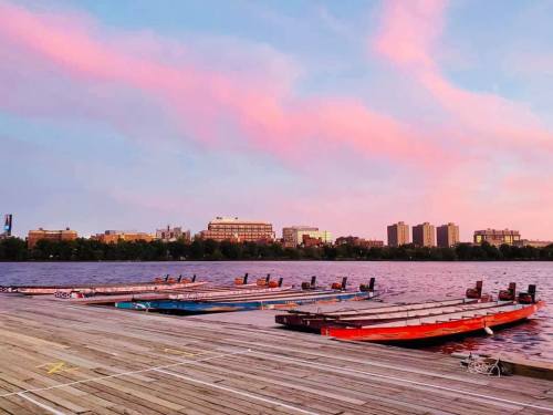 Boats lined up on a wooden dock at sunset, with a colorful sky and city skyline in the background.