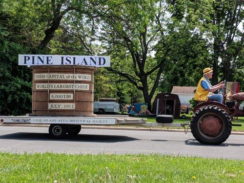 A tractor pulls a float displaying "Pine Island: Cheese Capital of the World" with a large cheese replica and historical details.