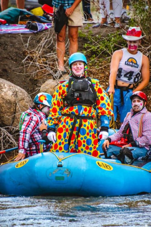 A group of clowns in colorful outfits on a blue raft, preparing for a river adventure.