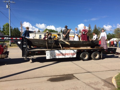 A group of people in historical costumes stands on a trailer with a large canoe, promoting the Grand Encampment Museum.