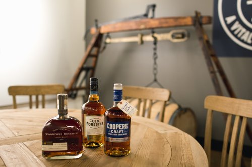 Three whiskey bottles on a wooden table, with a vintage distillery backdrop featuring a large wooden structure.