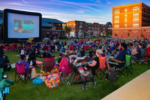 A large crowd sits on lawn chairs watching a movie on an inflatable screen in a park during sunset.