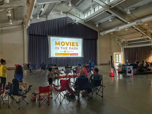 A community event with people sitting in chairs watching a movie on a large screen indoors.