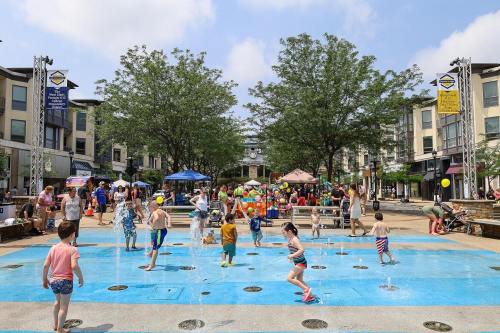 Children play in a splash pad surrounded by families and trees, with colorful tents and a lively atmosphere.