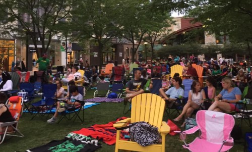 A crowd of people sitting on lawn chairs and blankets in a park, surrounded by trees and buildings, enjoying an outdoor event.