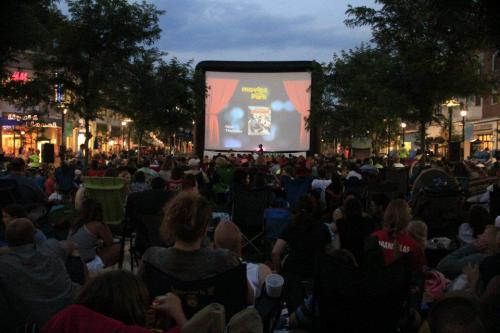 A large crowd sits on lawn chairs watching a movie on an outdoor screen at dusk, surrounded by trees and buildings.