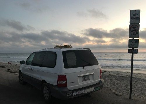 A white minivan parked near a beach at sunset, with a "No Parking" sign in the background.