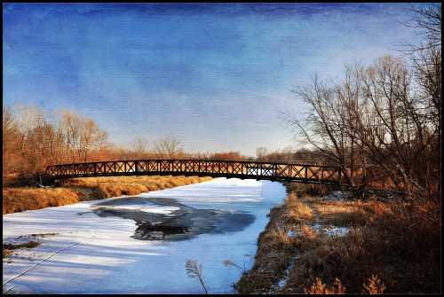 A wooden bridge spans a frozen river, surrounded by bare trees and a clear blue sky.