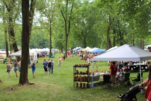 A vibrant outdoor market with tents, people browsing, and green trees in a park setting.