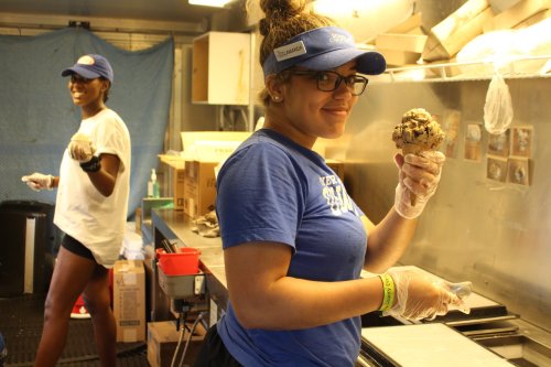 Two people in an ice cream shop; one holds a cone while the other prepares ice cream in the background.