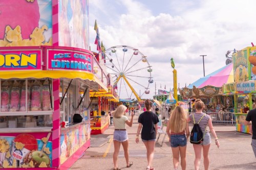 A group of friends walks through a colorful fairground with food stalls and a Ferris wheel in the background.