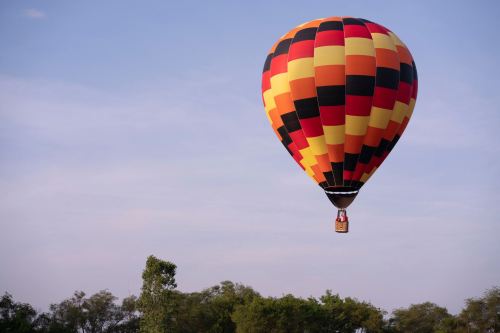A colorful hot air balloon with a checkered pattern floats above green trees against a clear blue sky.
