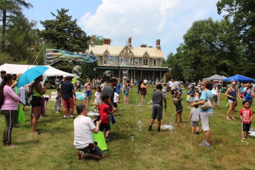 A lively outdoor event with families enjoying bubble activities on a sunny day, surrounded by tents and a historic house.