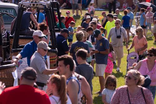 A crowded outdoor event with people interacting, enjoying activities, and a hot air balloon in the background.
