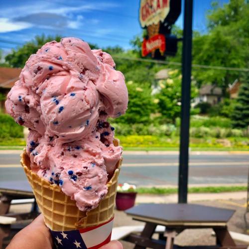 A hand holds a large cone of pink ice cream with blue sprinkles, set against a sunny outdoor scene.