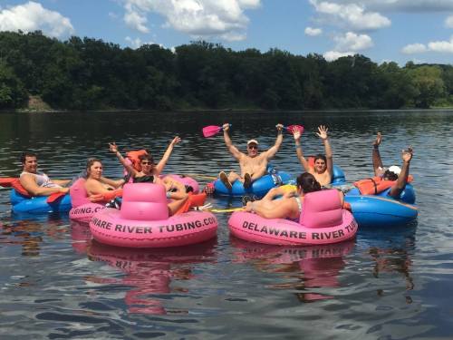 A group of people in colorful inner tubes on a river, smiling and waving, with trees and blue sky in the background.