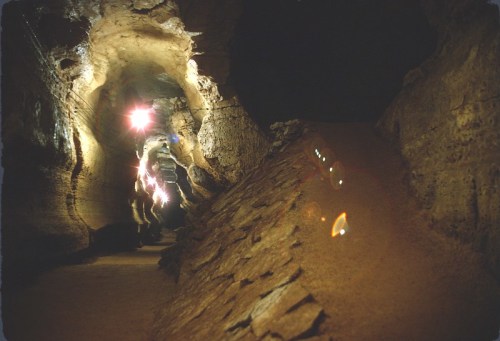 A dimly lit cave with rocky walls and a winding path, illuminated by colorful lights in the distance.