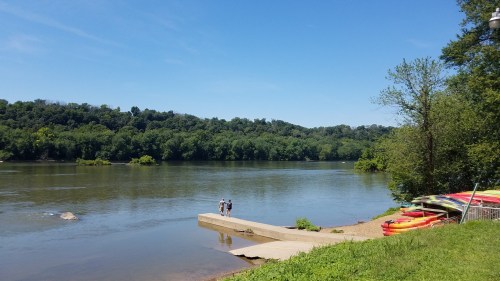Two people walk along a riverbank with lush green hills in the background and colorful kayaks on the shore.