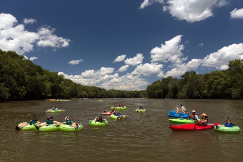 A group of people floating on colorful inner tubes in a river, surrounded by lush greenery and a blue sky with clouds.