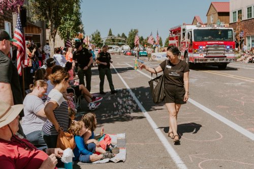 A parade scene with people sitting on the curb, a woman walking by with bubbles, and a fire truck in the background.