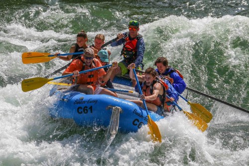 A group of six people in a blue raft navigating through whitewater rapids, paddling and enjoying the adventure.