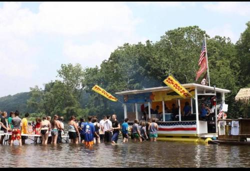 A crowd gathers around a floating food stand on a river, with trees and a blue sky in the background.