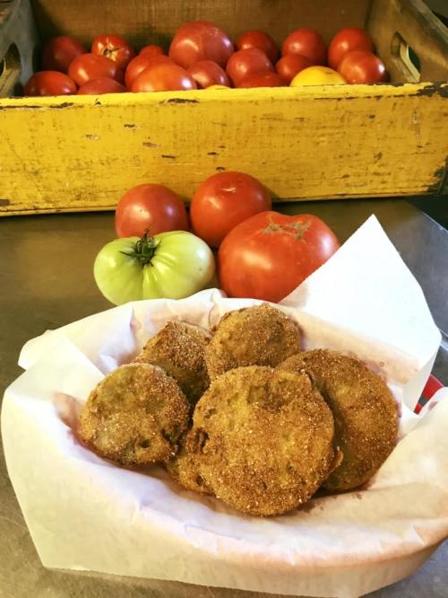 A basket of golden fried green tomatoes in front of fresh tomatoes and a yellow wooden crate.