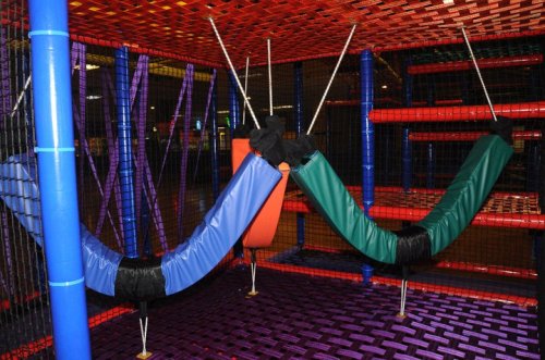 Colorful indoor play structure with hanging hammocks in blue, green, and orange, surrounded by netting and climbing areas.