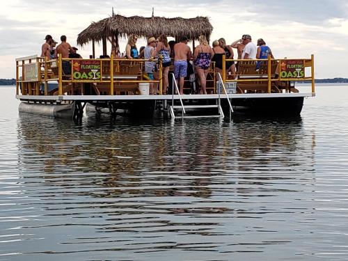 A group of people enjoying a floating bar with a thatched roof on calm water, reflecting the scene around it.