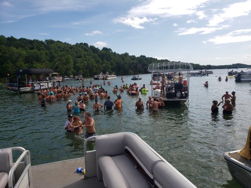 A crowded lake scene with people swimming and socializing, surrounded by boats and lush green hills in the background.