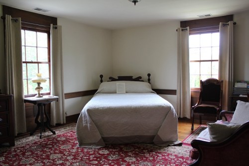 A cozy bedroom featuring a bed, two windows, a lamp, and a patterned rug, with a chair and a small table.