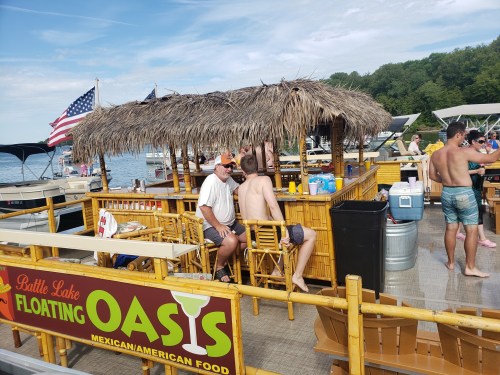 A lively floating bar with a thatched roof on a lake, surrounded by people enjoying the sun and water activities.