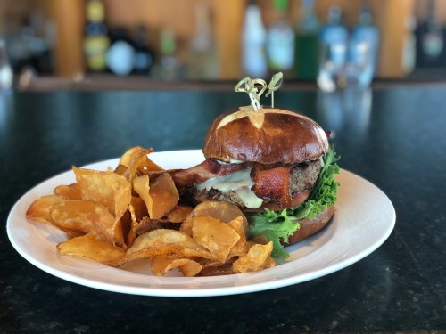 A juicy burger on a pretzel bun with lettuce, bacon, and a side of crispy potato chips on a white plate.
