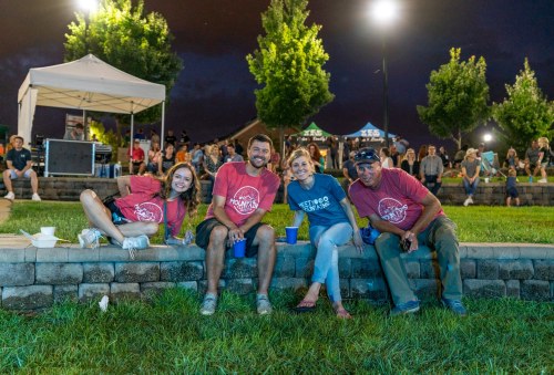 Four people sit on a stone wall in a park during an evening event, with trees and a crowd in the background.
