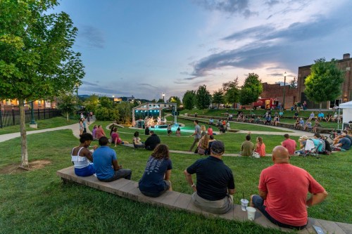 A crowd enjoys a live music performance in a park at dusk, with trees and buildings in the background.