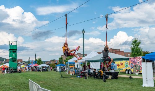 Two people ziplining across a grassy area with tents and a cloudy sky in the background.