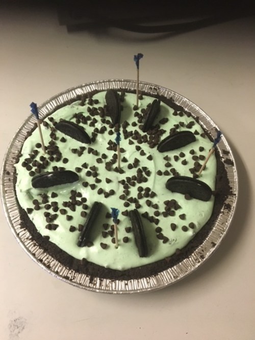 A mint green cake topped with crushed Oreos, chocolate chips, and blue candles, set in a silver pie dish.