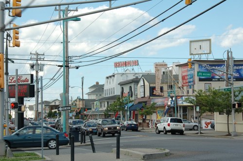 A busy street scene with shops, signs, and cars under a blue sky with clouds.