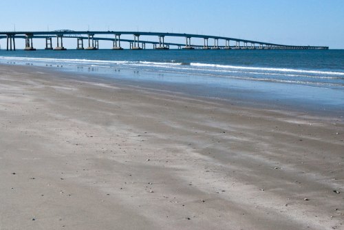 A long bridge stretches over calm waters, with a sandy beach in the foreground and clear blue skies above.