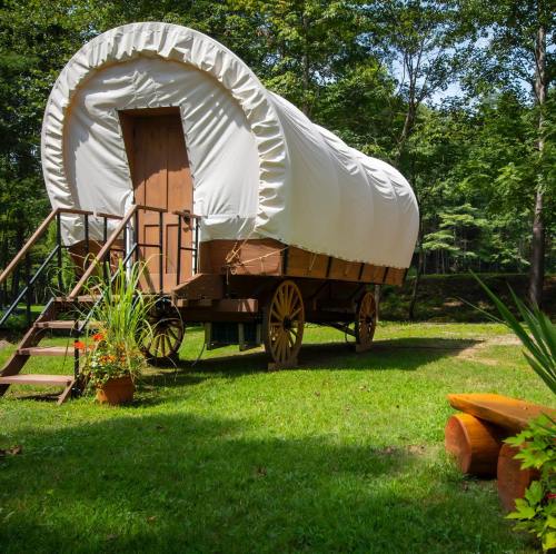 A covered wagon on wheels, set in a grassy area surrounded by trees, with steps leading up to the entrance.